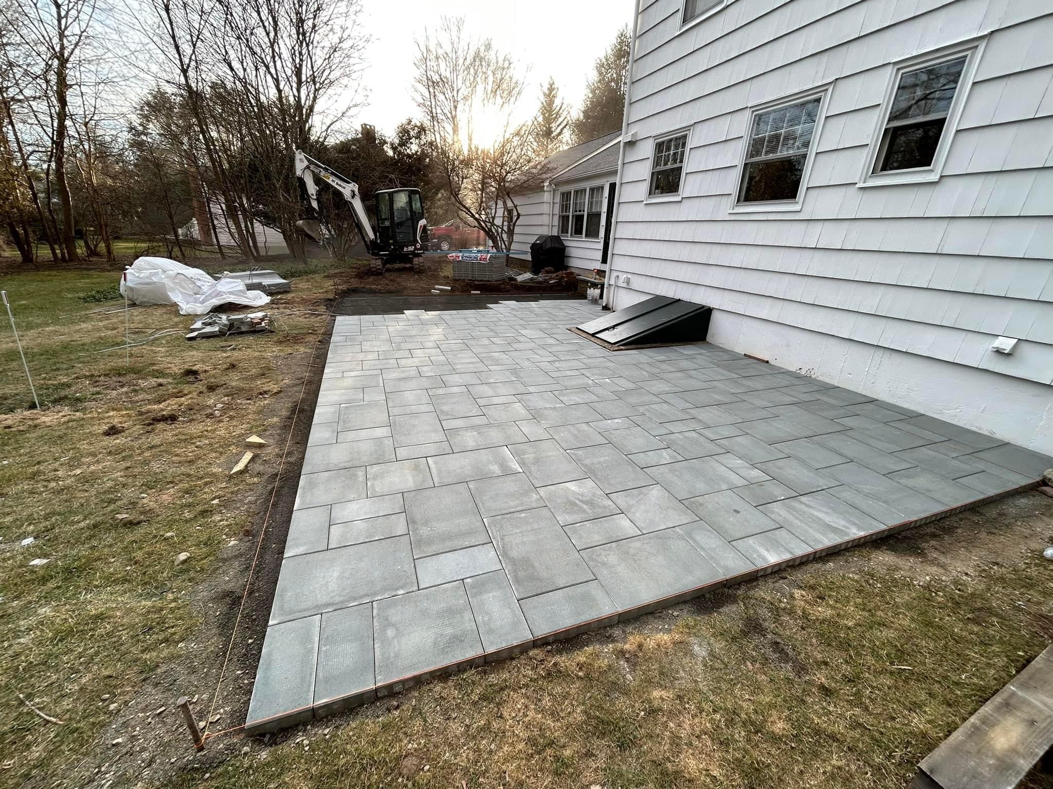 Paver patio in progress with mini excavator at white colonial home, showing construction phase