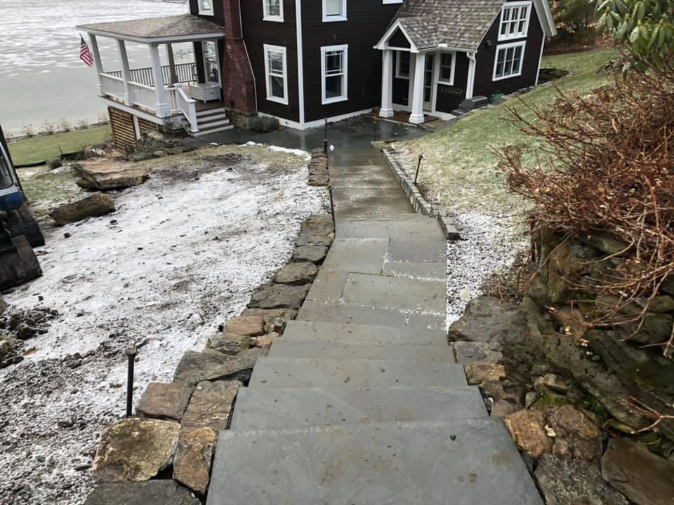 Bluestone walkway and slab steps descending hillside to dark colonial lakeside home with American flag
