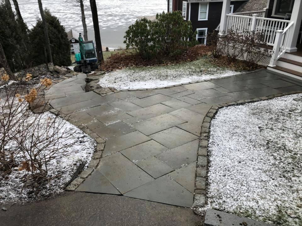 Bluestone herringbone walkway with cobblestone border curving past lakeside home with light snow