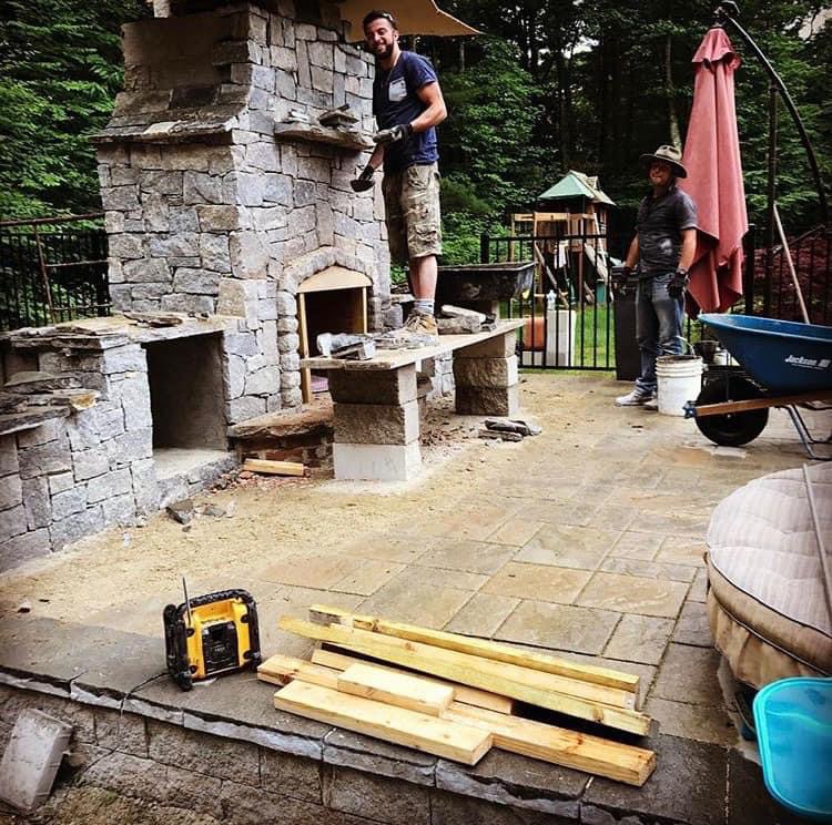 Crew building custom outdoor stone fireplace and pizza oven complex on flagstone patio, construction in progress