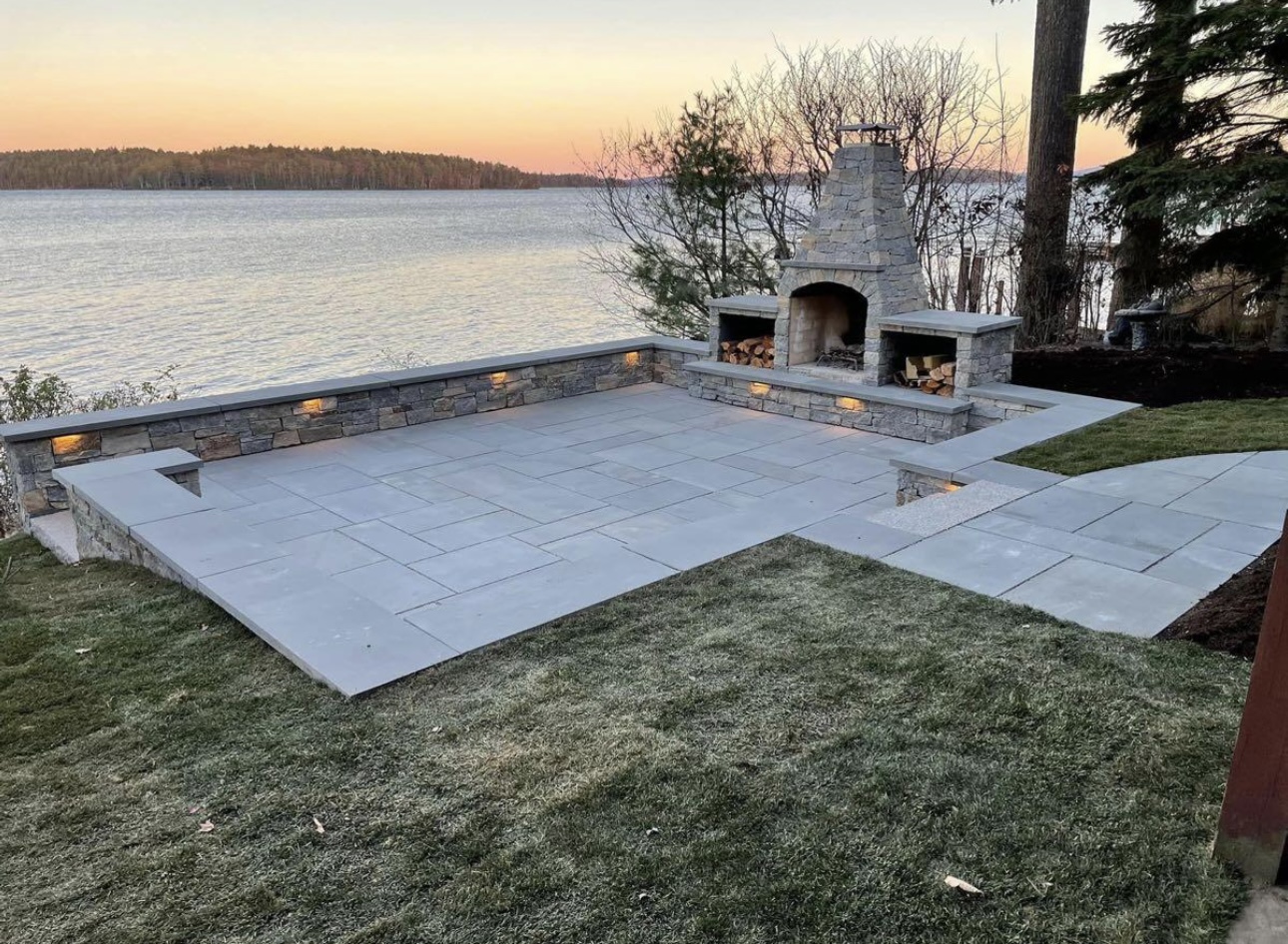 Lakeside patio with custom stone fireplace, bluestone pavers, and built-in wall lighting at sunset