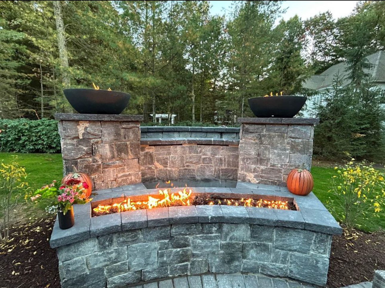 Custom stone fire feature with curved bench, dual fire bowls on pillars, and linear fire trough