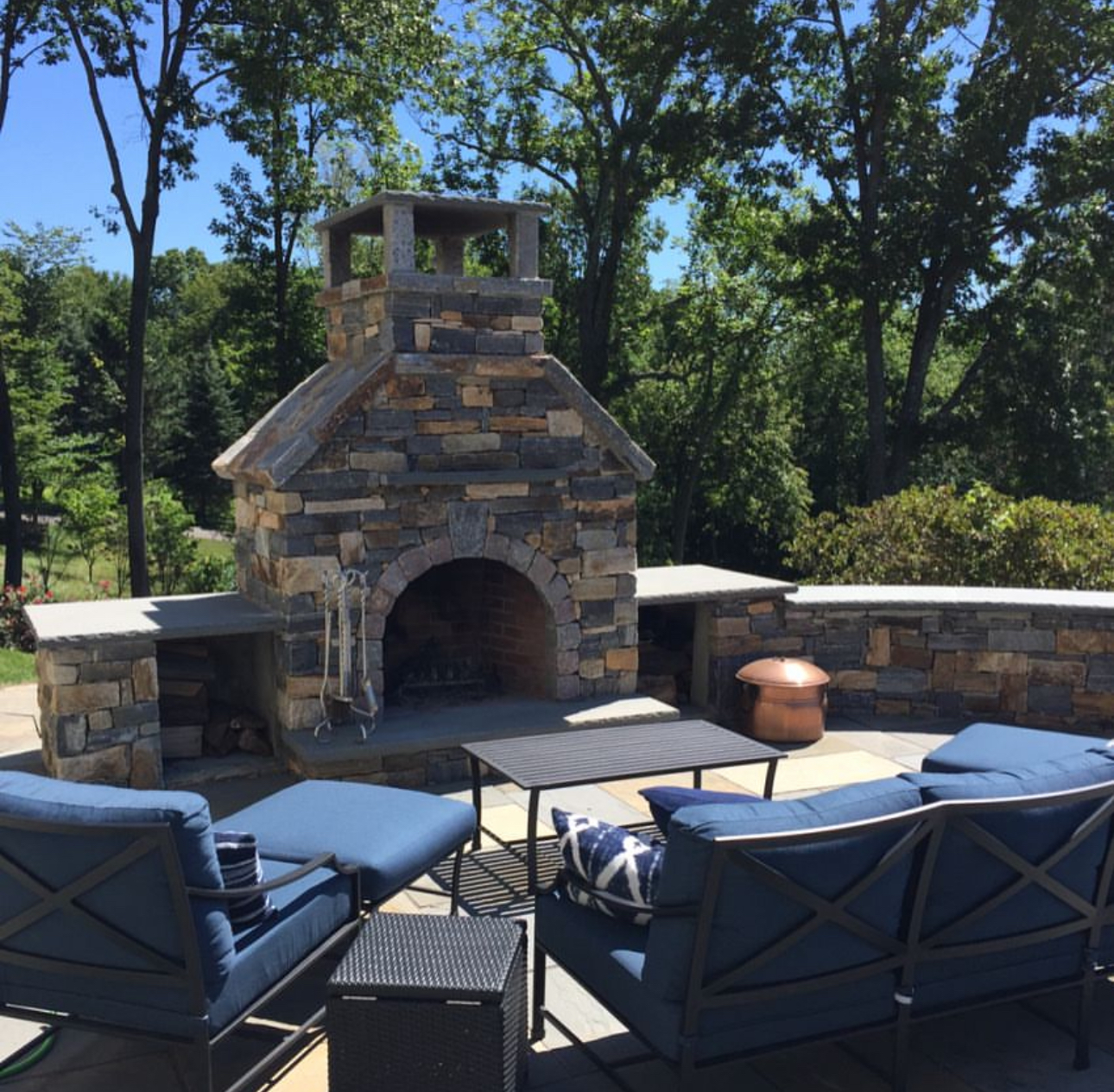Outdoor fieldstone fireplace with seating area, blue cushioned furniture, and copper accent pot