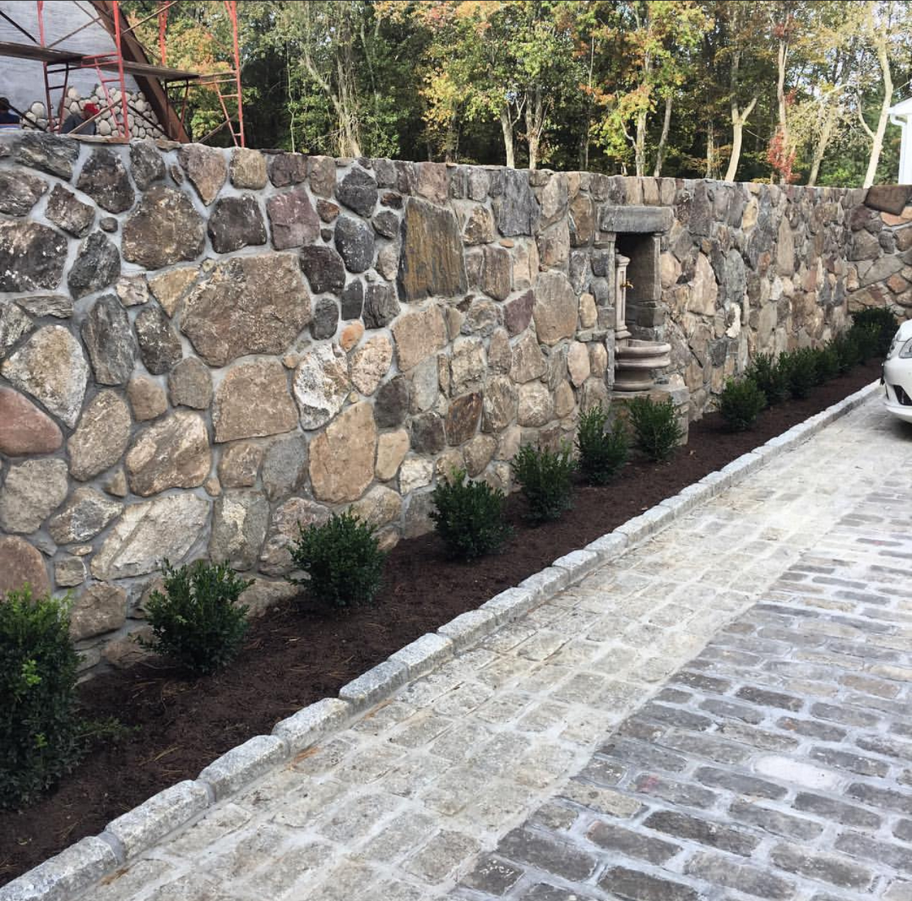 Massive fieldstone retaining wall with recessed niche, boxwood hedge plantings, and cobblestone driveway