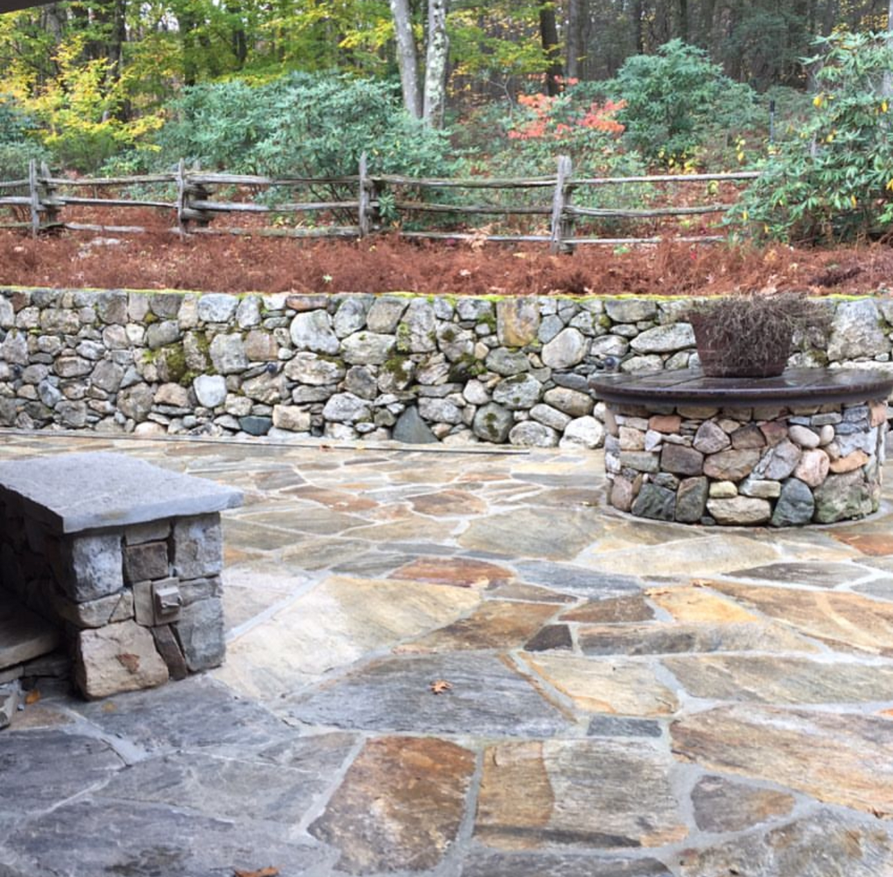 Flagstone patio with fieldstone fire pit, dry-stacked retaining wall, and split rail fence in autumn