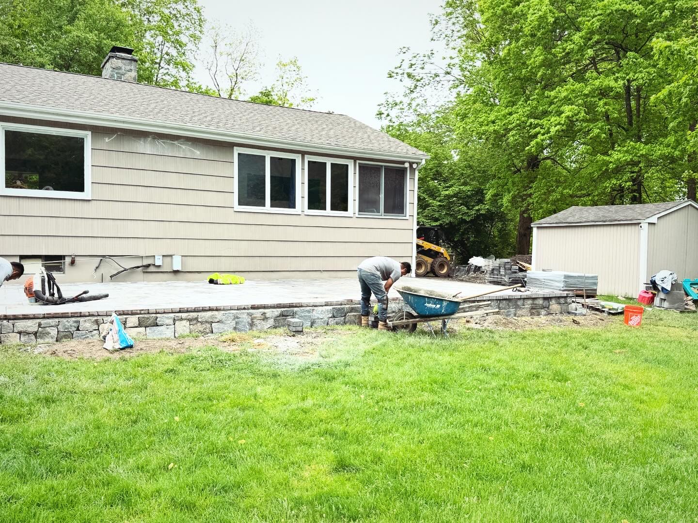 Raised paver patio under construction with stone foundation wall, workers laying pavers at residential home