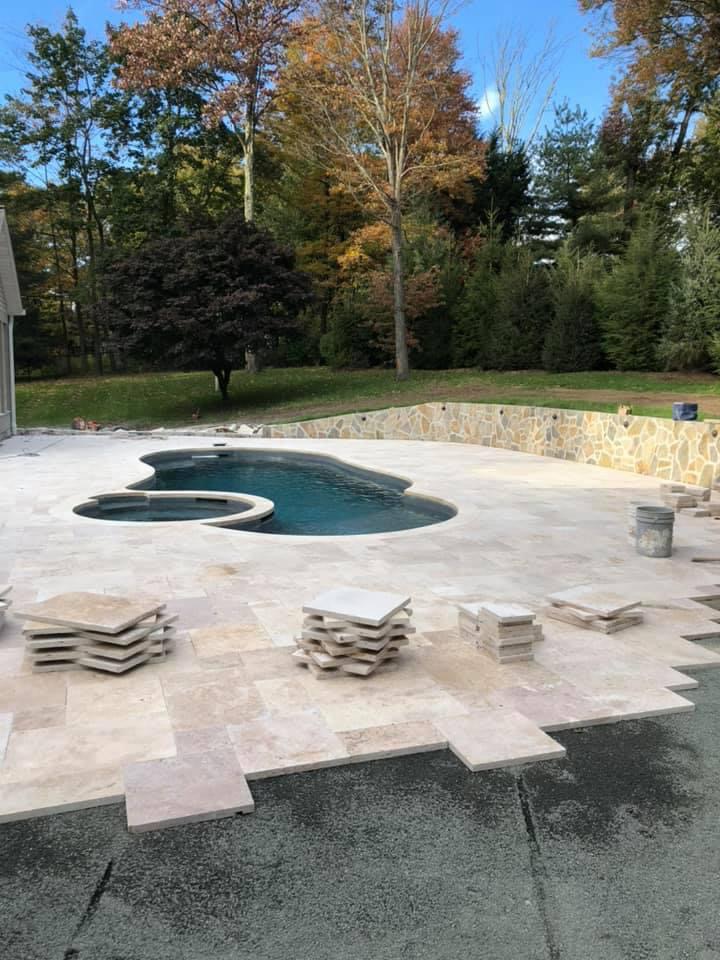 Travertine pool deck being installed around freeform pool with spa and fieldstone retaining wall in autumn